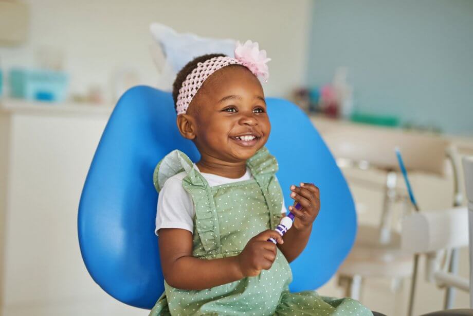female toddler child sitting in blue dental exam chair, holding a toothbrush