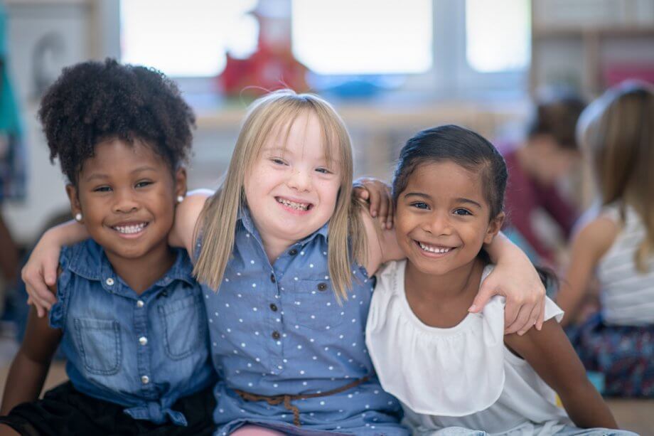 three kindergarten girls sitting on floor with arms around each other, smiling