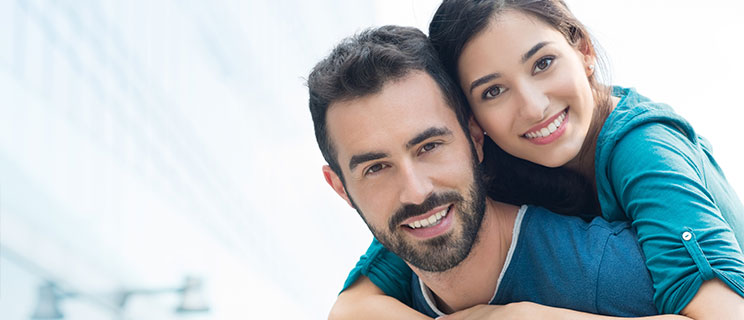 young smiling couple with woman's arms around man, looking into camera